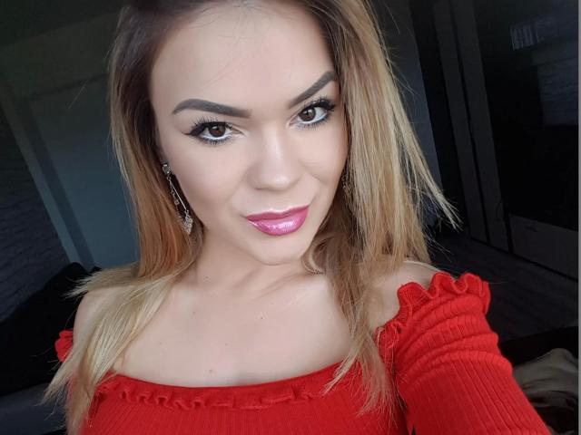 AngelSarah-hot with blonde hair, red off-shoulder top, pink lipstick, and eyeliner, showing a sexy smile, has cute tits, and is wearing dangling earrings, looking at the camera.