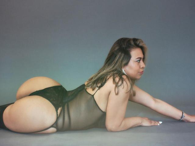 ValeriaFranco lying on her stomach, wearing a sheer black lace bodysuit, showcasing her big ass and curvy butt, with her head turned to the side, looking at the camera.