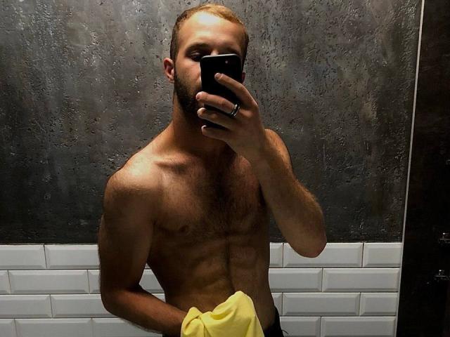 Shirtless LoveAndSexCouple with a muscular, hairy chest and visible abs, holding a yellow towel, taking a mirror selfie with a black phone, covering his face.
