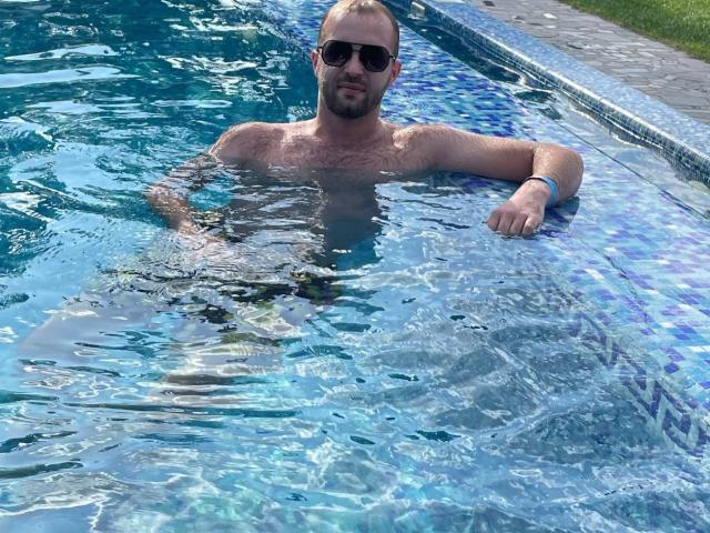 LoveAndSexCouple, shirtless, wearing black sunglasses, with a blue-tiled pool, arm resting on the edge, water up to chest, slight beard, wrist bracelet, looking relaxed.