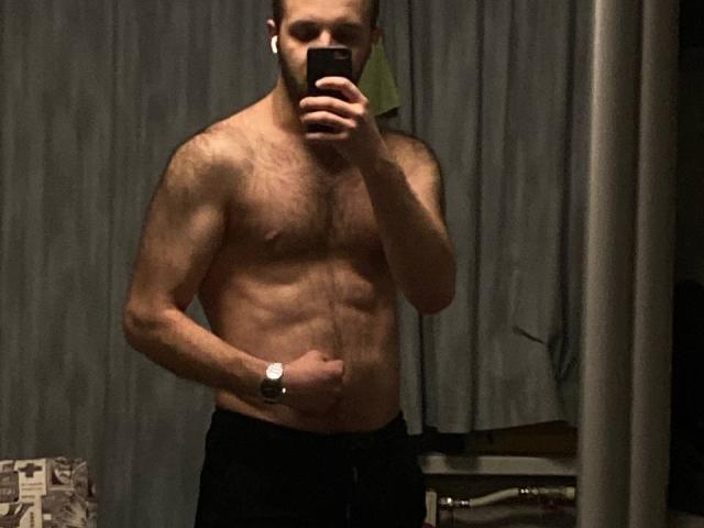 Shirtless LoveAndSexCouple with a muscular, hairy chest and visible abs, wearing black pants, holding a phone in front of their face, wearing a watch, in a dimly lit room.