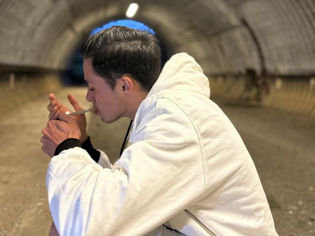 LoloHot with short dark hair, white hoodie, and black sleeves, sucking on a straw with hands near mouth, in a dimly lit tunnel with curved walls.