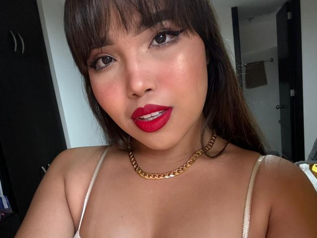 Heyly's face is close, red lipstick open, gold chain, brown hair, white strap top, looking at camera with sexy expression.