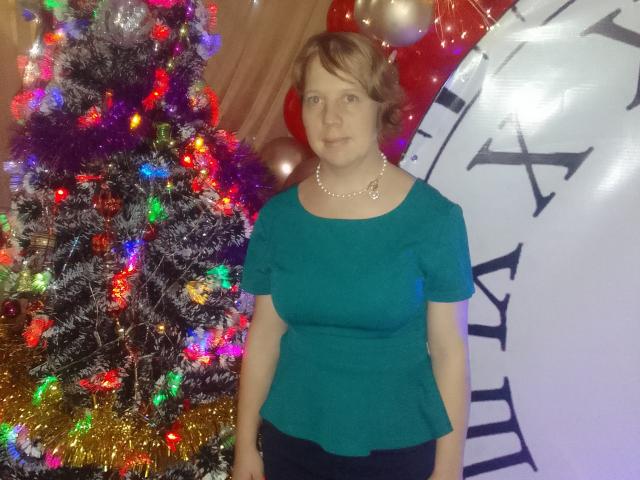 MariannaPope with short brown hair, wearing a teal shirt, pearl necklace, and dark pants, standing in front of a colorful Christmas tree with lights. Her tits are small.
