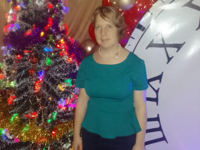MariannaPope stands in front of a Christmas tree with colorful lights, wearing a teal top, pearl necklace, and dark pants. She has short brown hair and a neutral expression.