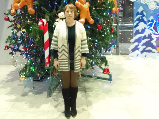 MariannaPope stands in front of a Christmas tree, wearing a black top, striped cardigan, brown tights, and black boots. She has short red hair and a neutral expression.