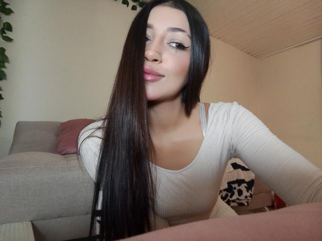 AnnaKhalo with long black hair, light pink lipstick, and a beige top, showing a slight smile, sitting on a couch with a pink pillow and green leaf garland.