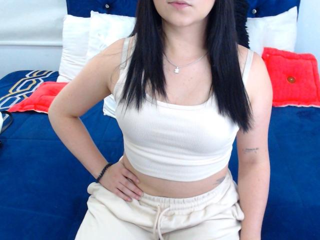 Kkatalina with black hair, white tank top, and beige pants, sitting on a blue bed. Visible small tattoo on right arm, white pillow, red pillow, and blue bed.