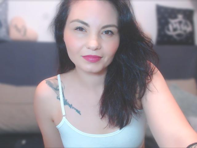 SexySunSex with dark hair, pink lipstick, white tank top, tattoo on left shoulder, slight smile, selfie, medium breasts, light skin, blurred background, soft lighting, relaxed expression.
