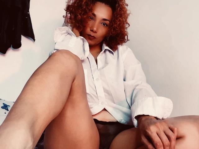 AmberDemons with curly red hair, wearing an unbuttoned white shirt and black panties, sitting with legs spread, showing her pussy, in a relaxed, slightly provocative pose.