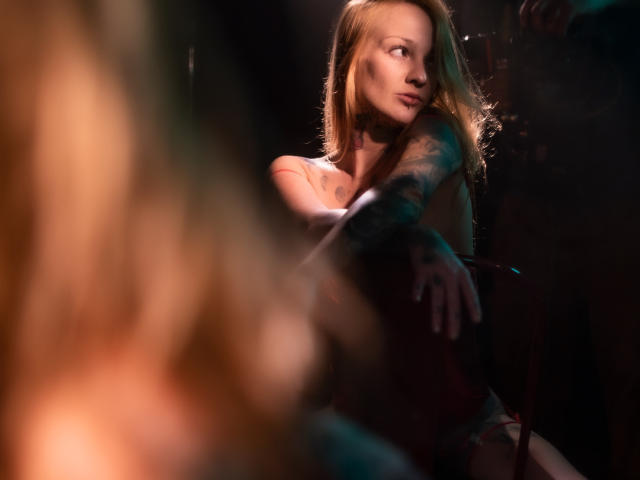 ScorpionLadyLuck with long blonde hair, tattoos on arm, topless, looking to the side, blurred foreground, dim lighting, sensual, soft shadows, slight smile, subtle makeup, warm tones.