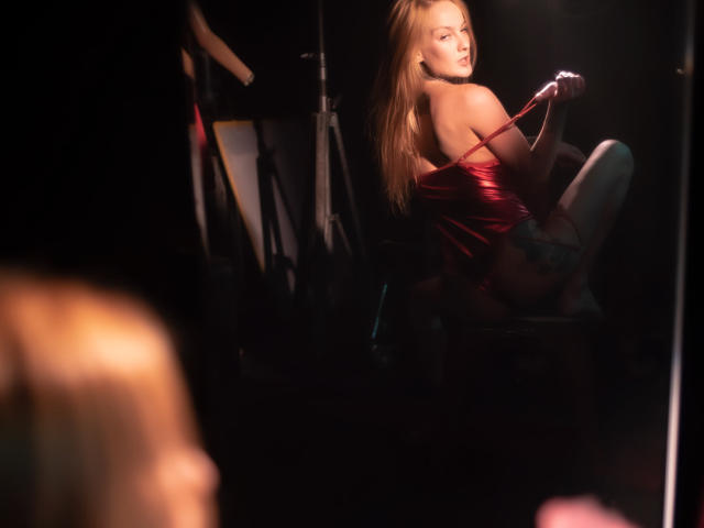 ScorpionLadyLuck with long blonde hair, wearing a red shiny dress, sitting on a stool, holding a microphone, looking over her shoulder, dimly lit, sexy, confident, showing her ass.