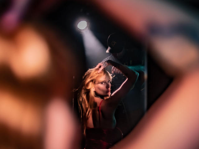 ScorpionLadyLuck with blonde hair, wearing a red dress, has her back to the camera, arms raised, under a spotlight, with a man in a cap partially visible.