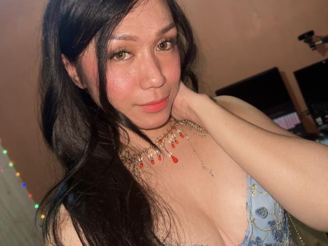TsJade with long black hair, pink cheeks, wearing a blue lace top with gold chains and red beads, touching her neck, green eyes, slight smile, showing her tits.