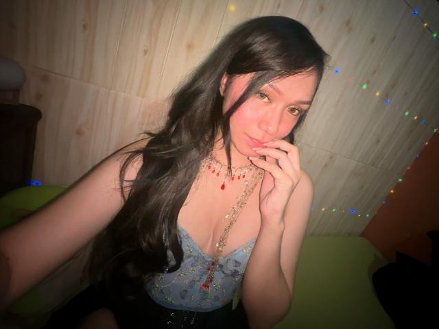 TsJade with long dark hair, blue lace top, and red necklace, touching her chin, showing her small tits, sitting on a green chair, in a wooden room.