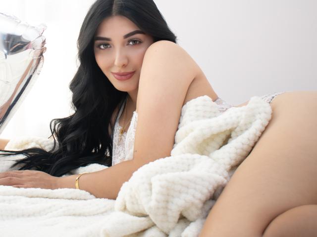 ArianaCortes with long black hair, lying on a white bed, wearing lace lingerie, butt in the air, white fluffy blanket, gold bracelet, smiling, shiny balloon on the left.