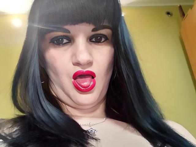 XLylyusX com cabelo azul escuro, batom vermelho e língua de fora, olhando para a câmera com a boca ligeiramente aberta, mostrando os dentes brancos. Blusa preta transparente, colar visível com design de teia de aranha..