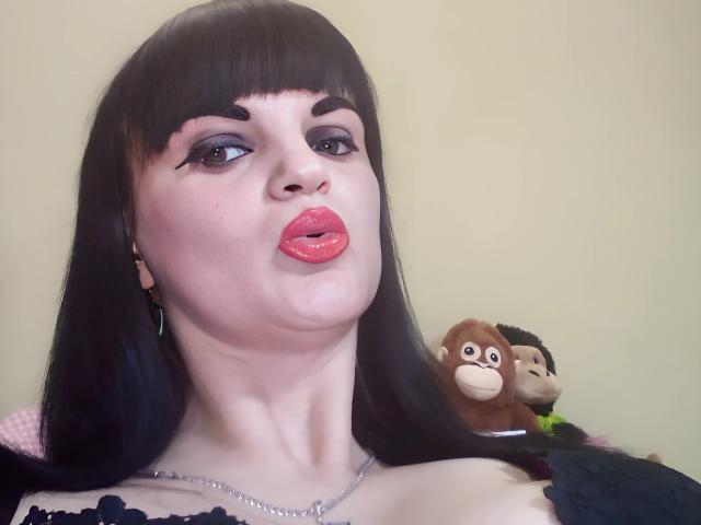 XLylyusX, com cabelo preto, batom rosa e delineador escuro, fazendo beicinho, usando um colar de prata, uma blusa de renda preta e com macacos de pelúcia ao fundo..