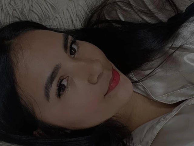 SofiaCopers has dark eyes, red lips, and black hair, lying on a bed with a white shirt. Her eyebrows are thick and she's smiling softly.