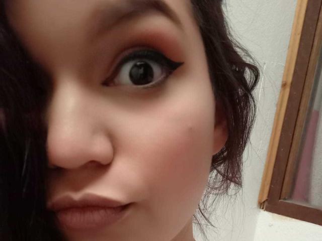 AriaLeenn's face, focusing on her eye with bold black eyeliner, pinkish-red eyeshadow, wavy dark hair, pale skin, and slightly parted lips. Her nose is centered.