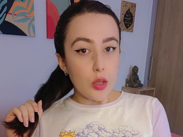 MabyLegrand with dark hair in a ponytail, pink lipstick, eyeliner, white shirt, holding hair, looking at camera, small earrings, Buddha statue in background, wooden door on right.