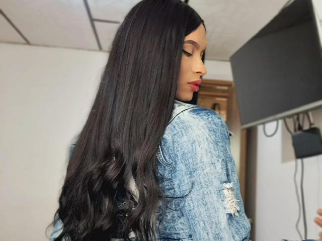 CoupleSensualHot avec de longs cheveux noirs et lisses, portant une veste en jean bleu déchirée, regardant sur le côté avec des lèvres rouges, dans une pièce avec une caméra noire sur la droite..
