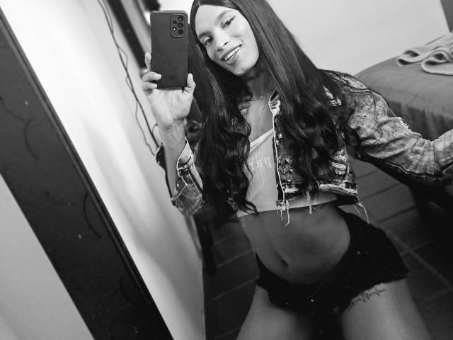 Black-and-white selfie of CoupleSensualHot with long dark hair, smiling, wearing a denim jacket, white crop top, and black shorts, holding a phone, showing a tattoo on her thigh.