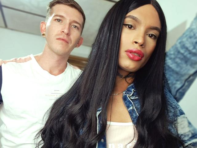 CoupleSensualHot, con lunghi capelli neri, rossetto rosso e giacca di jeans, è in posa di fronte a CoupleSensualHot, con capelli castani corti, maglietta bianca e piercing alle orecchie, in un selfie..