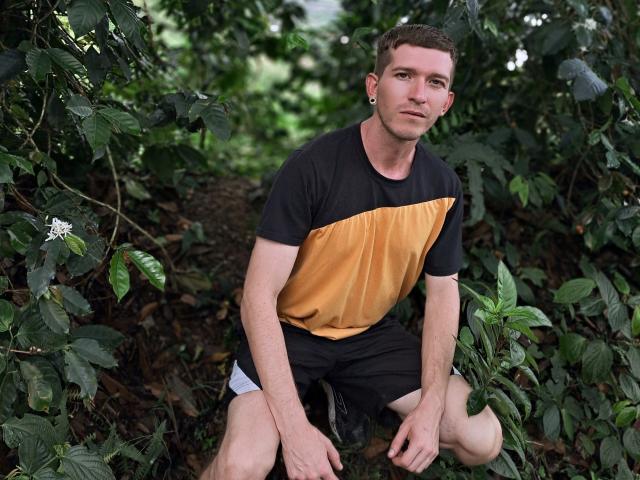 CoupleSensualHot, un homme musclé avec des cheveux bruns courts, un t-shirt noir et jaune, un short noir et des boucles d'oreilles, agenouillé dans un feuillage vert, regardant la caméra avec un léger sourire..