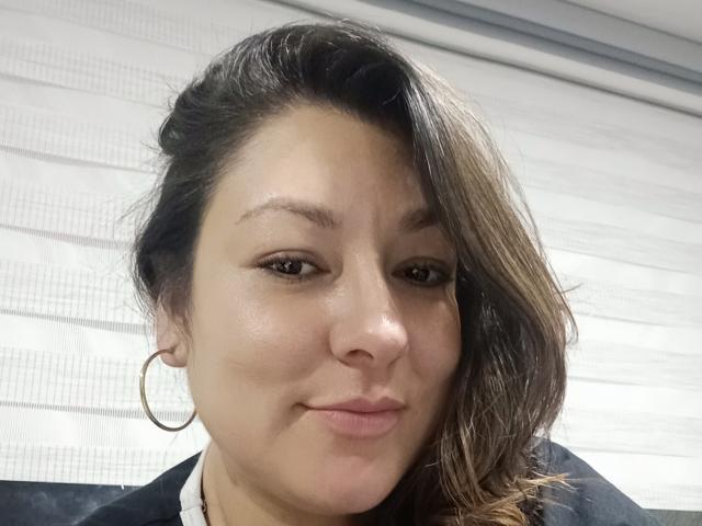 SakotoDika has light brown skin, brown eyes, and a slight smile with her nose slightly upturned. She has long brown hair, a gold hoop earring, and is wearing a black top.