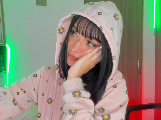 LindaWatsong, con cabello negro, flequillo y un piercing en la nariz, vestida con una sudadera con capucha rosa con estampado de margaritas, con el mentón apoyado en la mano, luces LED verdes y rojas en el fondo..