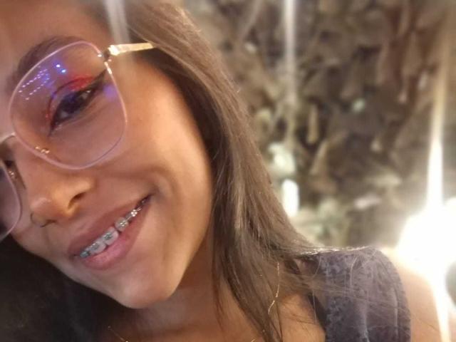 NoremCute with brown hair, wearing pink glasses, smiling at the camera, showing metallic braces on her teeth, in a soft-lit, blurry background.