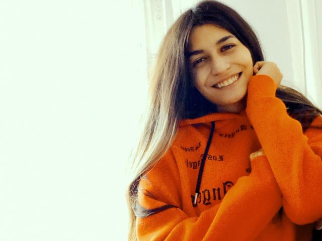 AnonimaKis with long brown hair, smiling, wearing an orange hoodie, hand near face, bright light from left, casual, cute, soft texture, warm color, relaxed, happy expression.