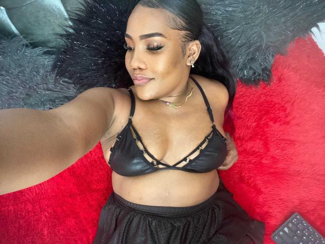SamiiaSmith with dark skin, black bra with strappy design, showing her small tits, lying on red and black furry textures, taking a selfie with a slight smile.