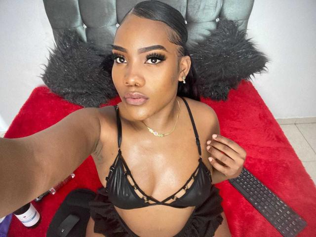 SamiiaSmith with dark skin, wearing a black strappy bikini top, gold necklace, and black fur pillow, taking a selfie, showing her tits and a remote.