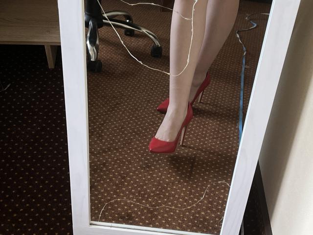 Les jambes de Valissia, chaussées de talons rouges, se reflètent dans un miroir, avec des câbles blancs sur le tapis. Elle porte des bas blancs transparents. Ses talons sont pointus et brillants..