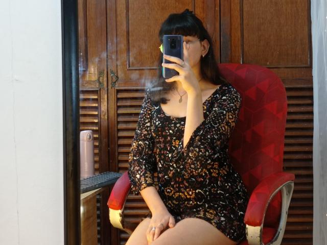 NatalyCloud with dark hair and bangs, wearing a black floral dress, sitting on a red chair, taking a mirror selfie with a phone, showing her breath.