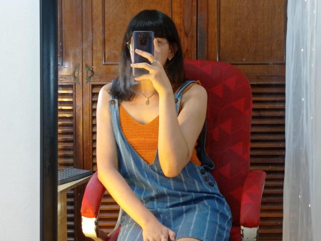 NatalyCloud with shoulder-length black hair, orange top, and blue striped overalls, sitting in a red chair, taking a mirror selfie, showing her tits and holding a phone.