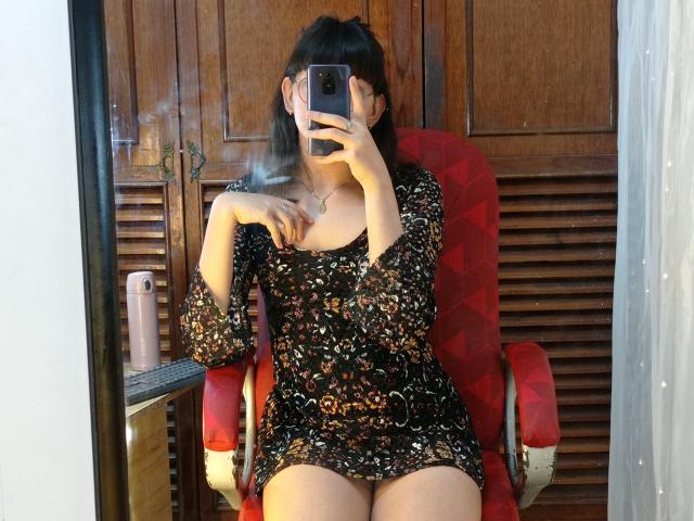 NatalyCloud with medium tits, sitting on a red chair, smoking, wearing a black floral dress, holding a phone, mirror selfie, wooden background, pink water bottle on left.