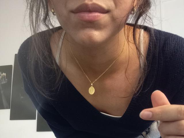 NatalyCloud's face, lower lip slightly up, wearing a gold necklace with a flat oval pendant, black sweater, and earrings, pointing with right index finger, brown hair.