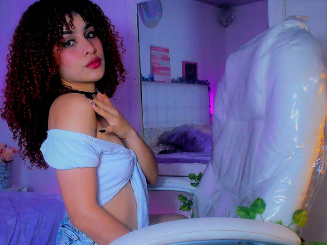 AmaiaEscotet with curly brown hair, wearing a blue off-shoulder top, touching her collarbone, sitting in a white chair, pink and purple lighting, looking at the camera.