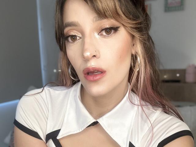 JaneRoa with fair skin, brown eyes, and pinkish lips, wearing a white top with black trim, hoop earrings, and brown hair with pink highlights, looking at the camera.