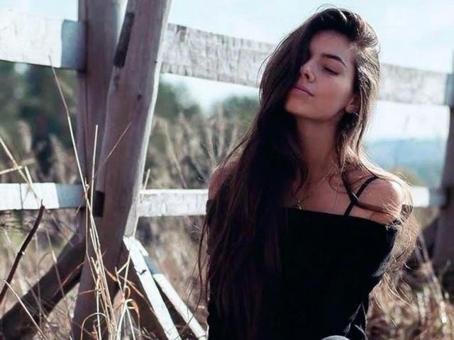 SensualMary-hot with long dark hair, eyes closed, wearing a black off-shoulder top, standing in front of a wooden fence, sunlight in her hair, looking sexy and calm.