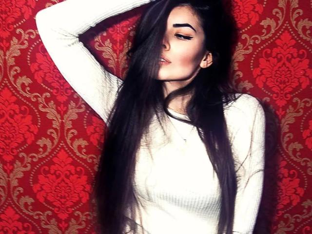 SensualMary-hot with long black hair, white ribbed sweater, raised arm, thick eyebrows, and slightly parted lips, showing off her tits and ass subtly. Red, floral-patterned background.