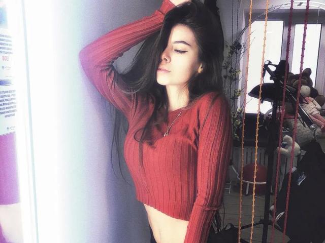 SensualMary-hot with long black hair, wearing a red ribbed crop top, standing with one hand in her hair, eyes closed, midriff exposed, in a softly lit room.
