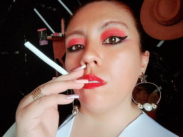 LexyDom with bright red eyeshadow, thick black eyeliner, red lipstick, holding a cigarette, wearing large silver earrings, and white shirt, with manicured nails touching her lips.