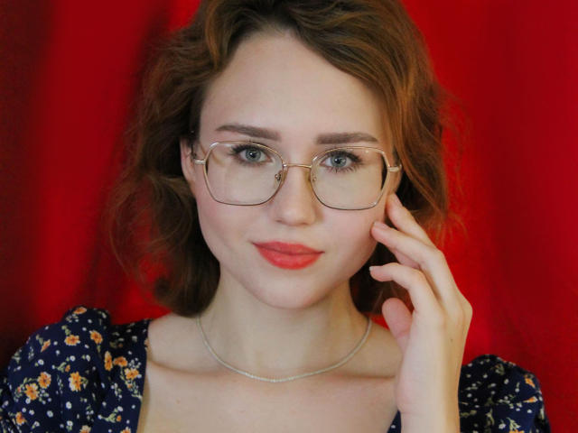 TeresaBirt with wavy brown hair, wearing glasses, red lipstick, and a floral dress, touching her cheek, pale skin, and a silver necklace, against a red background.