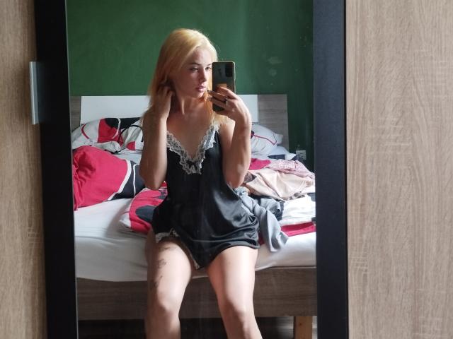 EvieEve with blonde hair, wearing a black lace-trimmed babydoll, sitting on a messy bed, taking a mirror selfie, showing her legs and a tattoo on her thigh.
