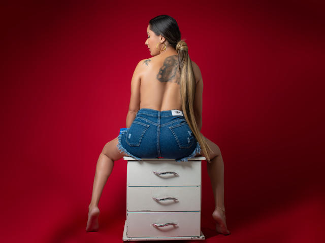 Topless CamilaBardot with long brown ponytail, denim shorts, sitting on white drawer, showing tattoo on right shoulder, red background, bare feet, back to camera, butt prominently.