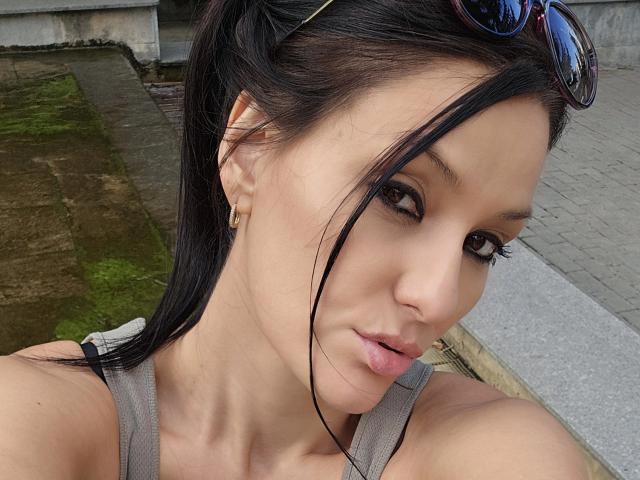 JoliDanielle with pouty lips, brown eyes, black hair in ponytail, wearing sunglasses on head, gold earrings, gray tank top, and light skin, sitting on a bench.